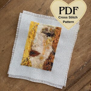 May include: A cross-stitch pattern on white fabric, depicting a face with yellow, brown, and beige hues. The text "PDF Cross Stitch Pattern" is on a heart-shaped tag. The fabric has a fringed edge.