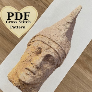 以下が含まれることがあります： ヘルメットをかぶった顔の石の彫刻の細部を描いたクロスステッチパターン。パターンは白いアイダクロスに印刷されています。