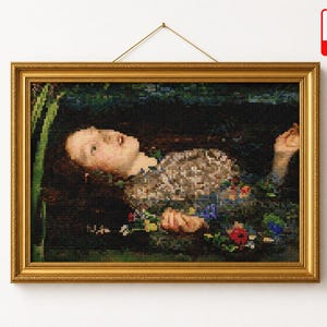May include: A cross-stitch embroidery of Ophelia floating in water, framed in a gold-colored rectangular frame. The image depicts a person with long dark hair, wearing a textured garment, surrounded by colorful flowers and green reeds.