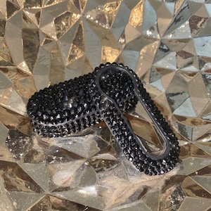 May include: A black key fob covered in black rhinestones. The fob is shaped like a car key with a keyhole and a button.