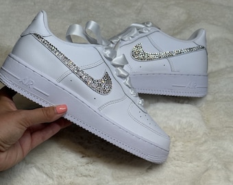 May include: White leather sneakers with a white satin ribbon tied around the ankle. The Nike swoosh is covered in clear rhinestones.