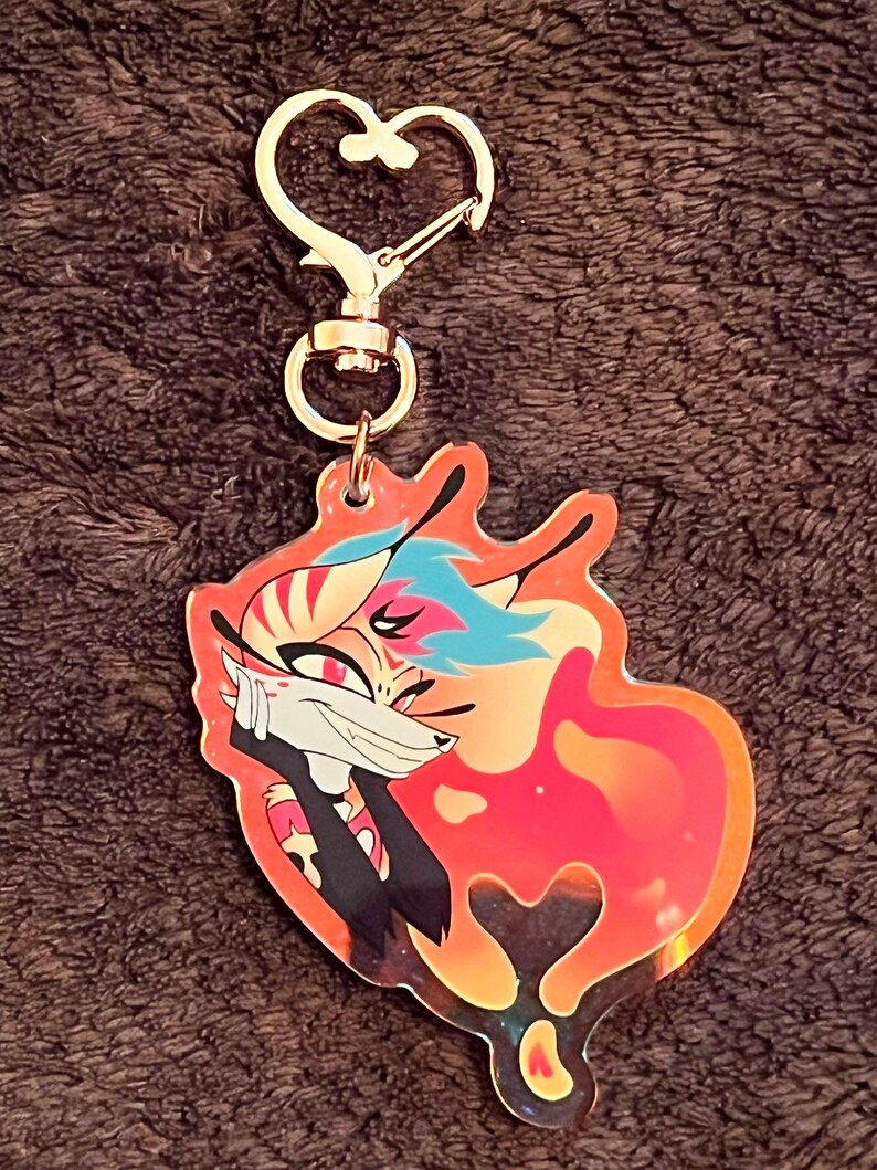 Puede incluir: Un llavero de coraz&oacute;n dorado con un personaje de zorro de dibujos animados colorido sobre un fondo rojo y naranja.