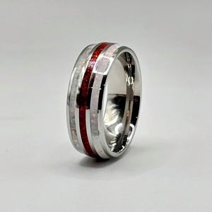 Puede incluir: Un anillo plateado pulido con una banda central de material incrustado rojo y blanco. El anillo tiene una superficie lisa y reflectante y un diseño de banda ancha. La incrustación roja contrasta con los elementos plateados y blancos.