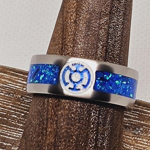 May include: A silver ring with a blue and white inlay. The inlay features a stylized symbol with a white center and blue outer ring. The ring is on a wooden surface.