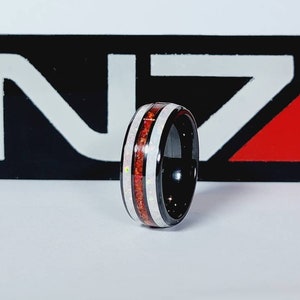 Puede incluir: Un anillo de carburo de tungsteno negro con incrustaciones rojas y blancas. El anillo tiene un acabado pulido y es una opción popular para la joyería masculina.