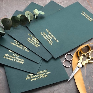 Könnte beinhalten: Ein Stapel dunkelgrüner Umschläge mit goldener Beschriftung, jeder mit einem anderen Namen und einer anderen Adresse. Die Umschläge liegen auf einer strukturierten Oberfläche mit einem goldenen Band und einer goldenen Schere.