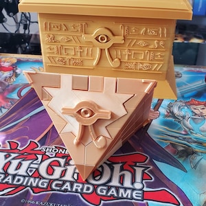 May include: A gold-colored 3D printed replica of the Millennium Puzzle from the Yu-Gi-Oh! Trading Card Game. The puzzle is a triangular prism with a golden eye symbol on the front. The puzzle is sitting on a Yu-Gi-Oh! Trading Card Game playmat.