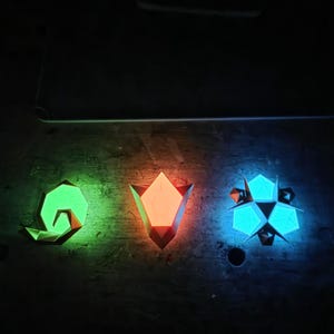 May include: Three geometric, glowing wall art pieces in green, orange, and blue. The shapes are a spiral, a tulip, and a starburst, each with a unique faceted design. The art is mounted on a dark, textured surface.