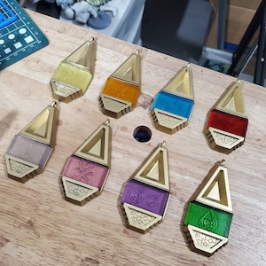 May include: A collection of eight pendants with a gold-colored metal frame and a colored, rectangular center. The colors include yellow, orange, blue, red, lavender, pink, purple, and green. Each pendant has a small loop at the top for a chain.