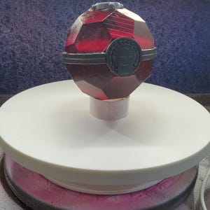Puede incluir: Una réplica de Poké Ball roja y negra con una base blanca. La Poké Ball es un objeto de colección popular para los fanáticos de la franquicia Pokémon.
