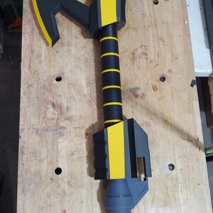 Puede incluir: Un hacha de utiler&iacute;a negra y amarilla con un mango gris y un logotipo en blanco y negro de una criatura en la parte superior de la cabeza del hacha.