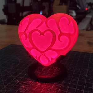 Peut inclure: Une lampe en forme de cœur rouge avec un design décoratif. La lampe est allumée et brille, projetant une lumière rouge chaude.