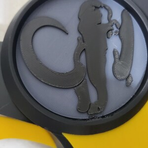 Puede incluir: Un bot&oacute;n circular negro y gris con una silueta negra en relieve y texturada de un mammut sobre un fondo gris. El bot&oacute;n est&aacute; unido a un objeto amarillo.