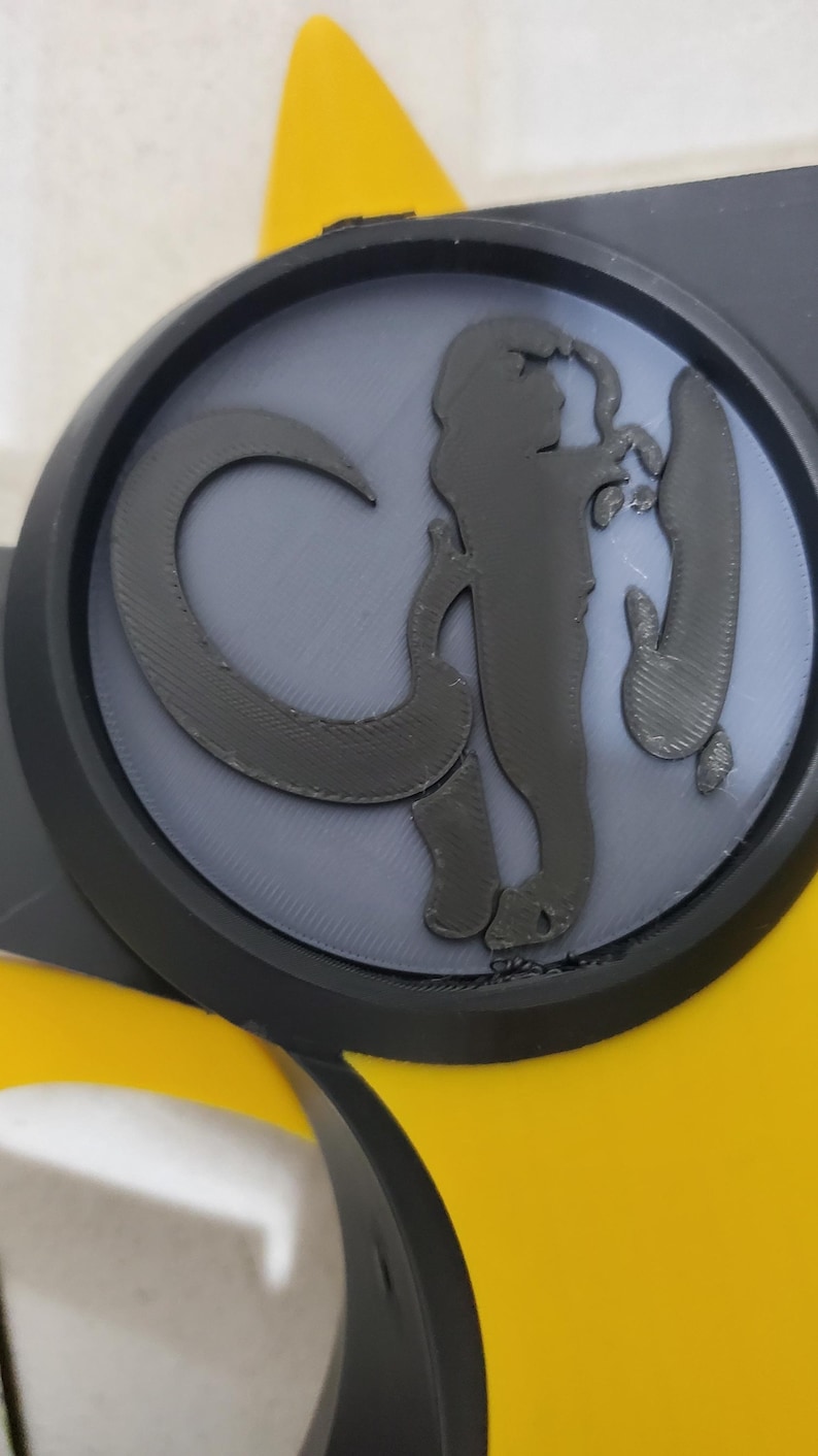 Puede incluir: Un bot&oacute;n circular negro y gris con una silueta negra en relieve y texturada de un mammut sobre un fondo gris. El bot&oacute;n est&aacute; unido a un objeto amarillo.