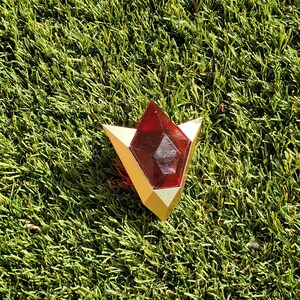May include: A red and gold triangular-shaped pendant with a red gem in the center. The pendant is lying on a green artificial grass surface.