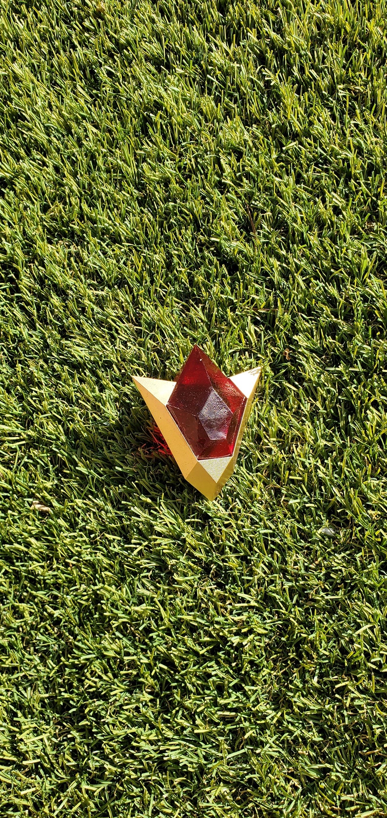 May include: A red and gold triangular-shaped pendant with a red gem in the center. The pendant is lying on a green artificial grass surface.