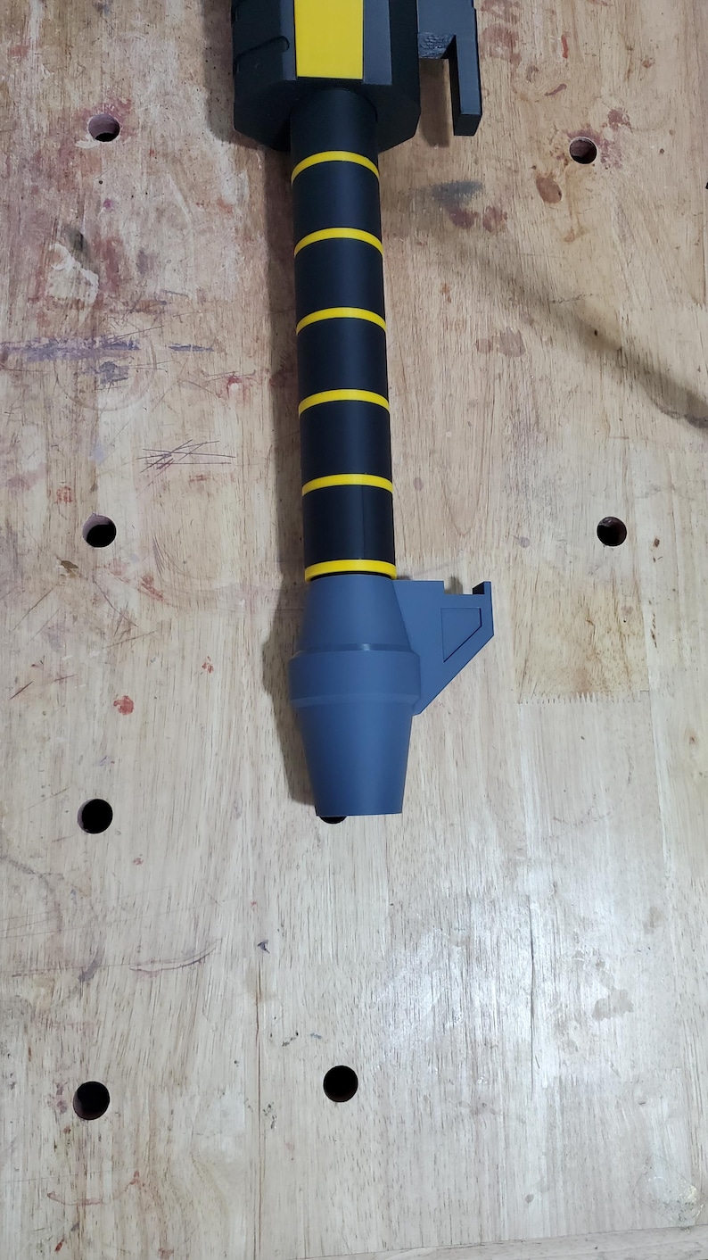 Puede incluir: Un mango de espada negro y amarillo con una punta de pl&aacute;stico gris. El mango tiene un agarre texturizado y un dise&ntilde;o a rayas negro y amarillo.