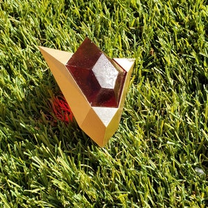 May include: A gold-colored, geometrically shaped pendant with a red gem in the center. The pendant is lying on a green artificial grass surface.