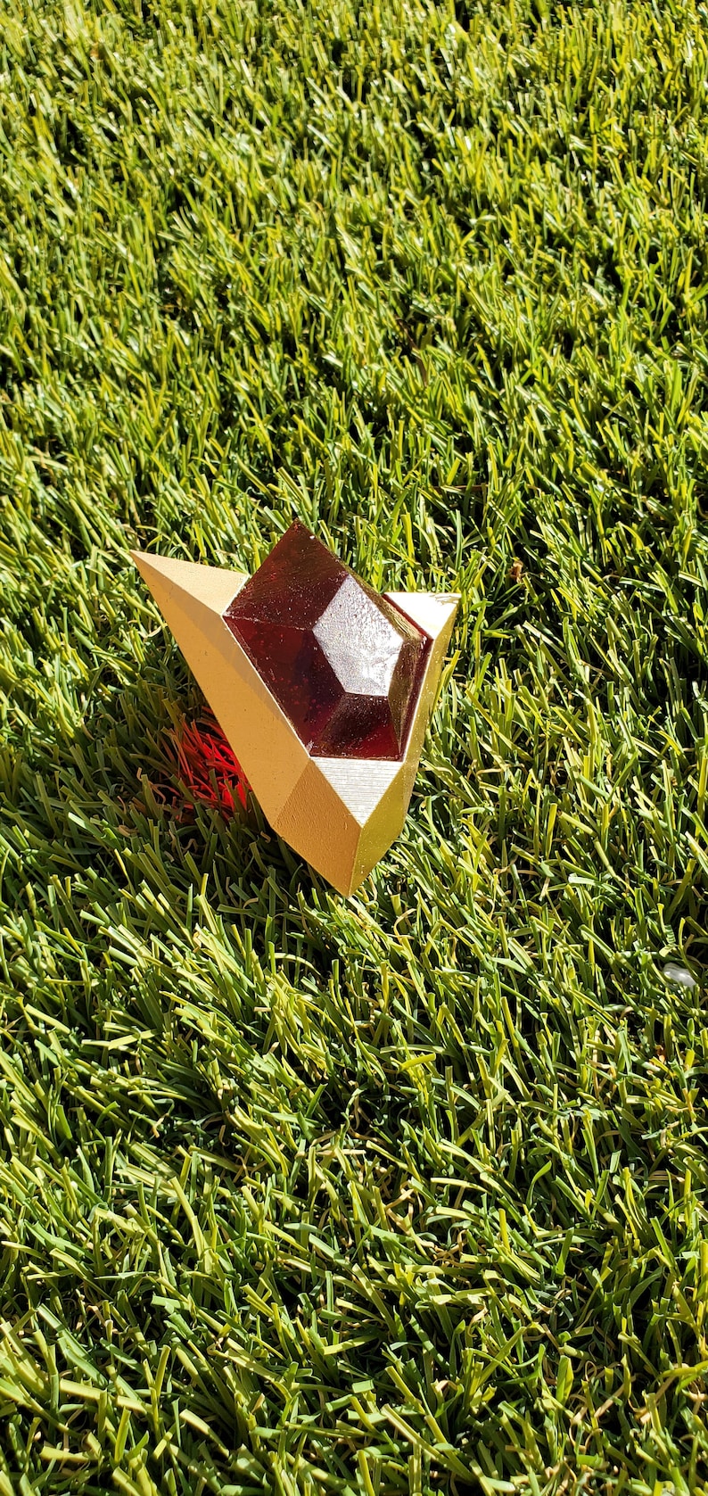 May include: A gold-colored, geometrically shaped pendant with a red gem in the center. The pendant is lying on a green artificial grass surface.
