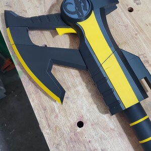 Puede incluir: Un hacha negra y amarilla con un dise&ntilde;o estilizado de cabeza de animal en el mango. El hacha tiene una hoja afilada y un mango texturizado.