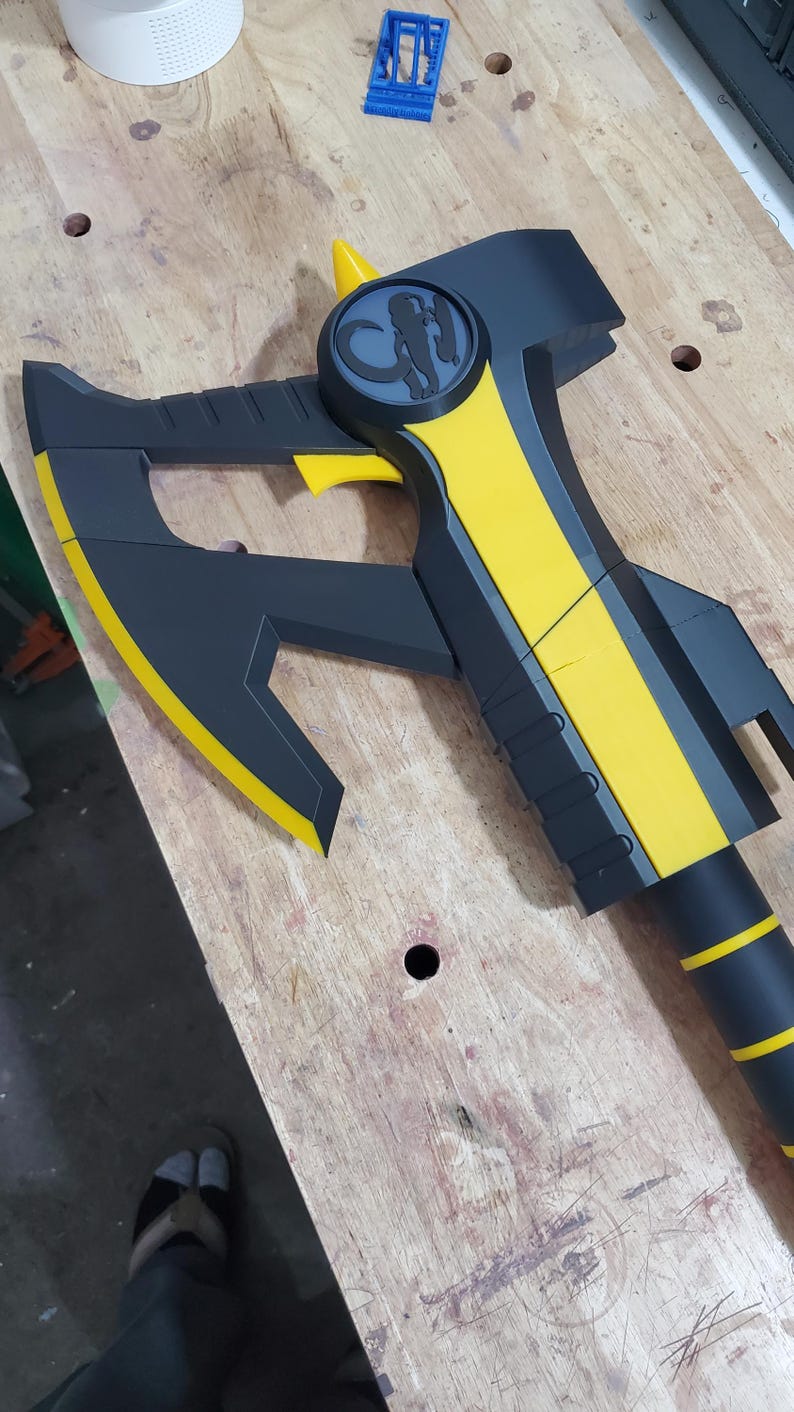 Puede incluir: Un hacha negra y amarilla con un dise&ntilde;o estilizado de cabeza de animal en el mango. El hacha tiene una hoja afilada y un mango texturizado.