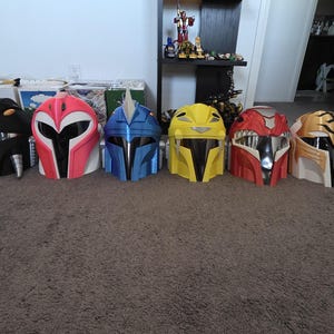 以下が含まれることがあります： 様々な色の7つのパワーレンジャーのヘルメットのコレクション。各ヘルメットは、緑、黒、ピンク、青、黄、赤、金など、独特のデザインと配色を持っています。ヘルメットはカーペットの上に並んでいます。