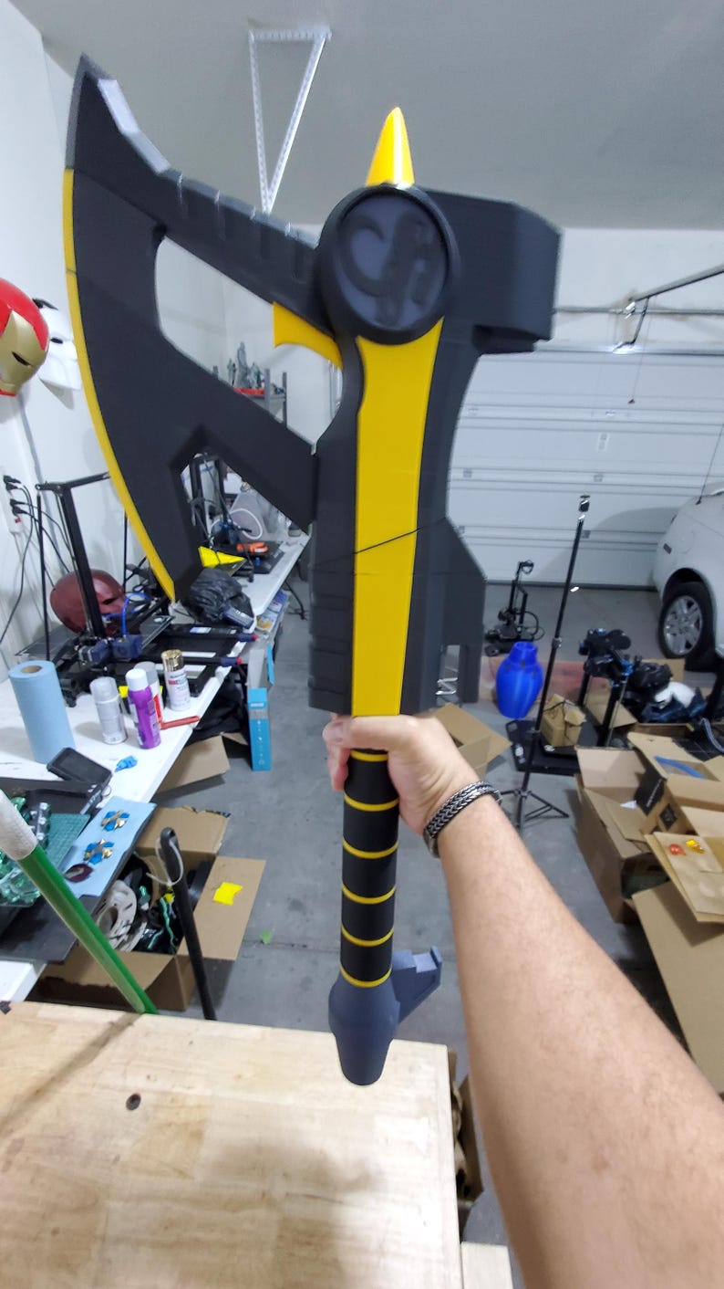 Puede incluir: Un hacha negra y amarilla con un mango texturizado. El hacha tiene una hoja afilada y una punta puntiaguda. El hacha est&aacute; sostenida por la mano de una persona.