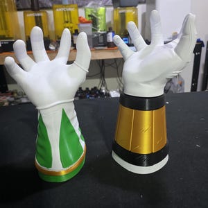 Op de afbeelding: Twee witte handschoenen met open handpalmen en gedetailleerde vingers en handen. Eén handschoen heeft een groene en witte manchet, de andere een gouden en zwarte manchet. De handschoenen lijken 3D-geprint, waarschijnlijk cosplay- of kostuumaccessoires.