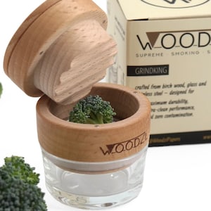Könnte beinhalten: Eine Kräutermühle aus Holz und Glas mit abgenommenem Deckel, in der ein kleines Stück Brokkoli liegt. Die Mühle trägt den Markennamen WOOD.ZL. Daneben steht eine beige Pappschachtel mit der Aufschrift WOOD.ZL GRINDKING und Produkttext.