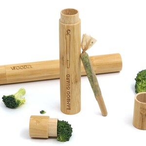 Può includere: Un tubo di stoccaggio in legno con la scritta "BAMBOO GUARD" incisa sul lato. Il tubo è aperto e contiene una sostanza verde arrotolata. Il tubo è appoggiato su una superficie bianca con alcuni pezzi di broccoli.