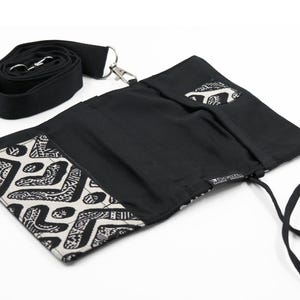 Könnte beinhalten: Eine schwarze Stofftasche mit einer weiß-schwarz geometrisch gemusterten Klappe, ausgestattet mit einer Innentasche, einem abnehmbaren schwarzen Riemen mit silbernem Metallverschluss und einem kleinen schwarzen Stoffetikett mit weißem Text "RAW SUPREME - SMOKING".