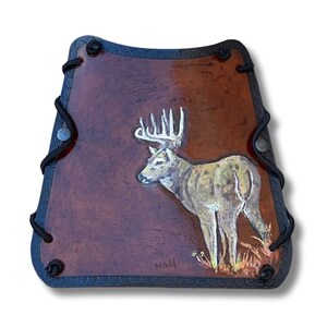 May include: A brown leather archery armguard with a painted deer image. The deer is tan and white with large antlers, standing in a field of grass. The armguard has black trim and black cord lacing.