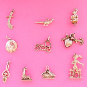 May include: A collection of gold-toned vintage charms, including a sword, bird, seated figure, locket, car, heart with cross and anchor, musical note, church, house, and a woman with a fence, against a pink background.