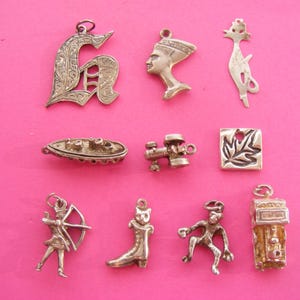 以下が含まれることがあります： ピンク色の背景に、銀色のチャームのコレクション。チャームには、装飾的な文字「G」、ファラオの胸像、猫、ボート、カメラ、葉、射手、ブーツ、踊る人物、建物が含まれています。
