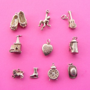 May include: A collection of silver charms on a pink background. Charms include ballet slippers, a monkey, a guitar, a church, a heart, a lantern, a lion, an ice skate, a clock, and a cowboy hat.