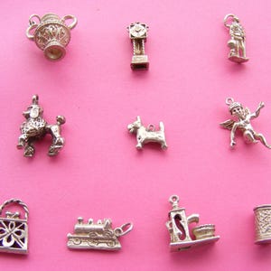 May include: A collection of silver-toned charms, including a poodle, a dog, a cherub, a teapot, a clock, a soldier, a purse, a train, a coffee maker, and a mug. The charms are displayed against a pink background.