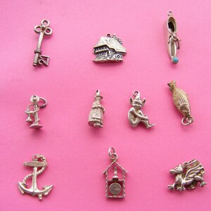 May include: A collection of silver charms on a pink background. The charms include a key, a house, a shoe with a turquoise bead, a lamp, a woman, a seated elf, a fish, an anchor, a clock, and a dragon.
