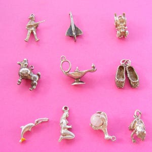 May include: A collection of silver-toned charms on a pink background. The charms include a soldier, a jet, a bear, a horse, a genie lamp, ballet slippers, a dolphin, a mermaid, a hand mirror with a pink stone, and a frog.