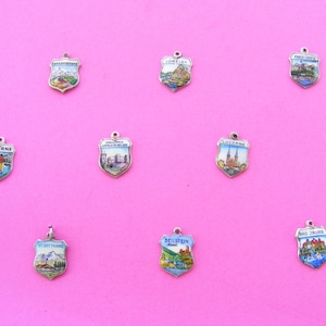 May include: A collection of vintage shield-shaped charms with colorful enamel designs. Each charm features a different European city or landmark, including Lucerne and Paris-Castle. The charms are displayed on a bright pink background.