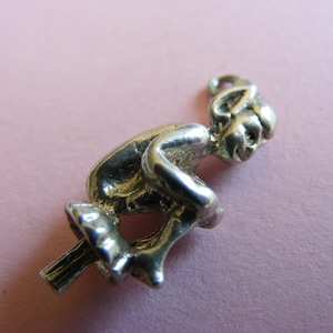 Puede incluir: Un charm plateado que representa una figura caprichosa montada en un palo. El charm presenta una carita sonriente y un diseño detallado. La figura está sentada en un palo, con las piernas extendidas. El charm es probablemente de metal y está sobre un fondo rosa.