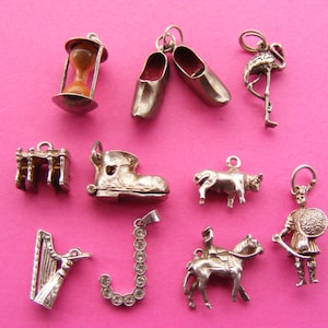 May include: A collection of silver charms on a pink background. Charms include an hourglass, shoes, a flamingo, a table, a boot, a bull, a harp, a letter J, a horse and rider, and a Roman soldier.