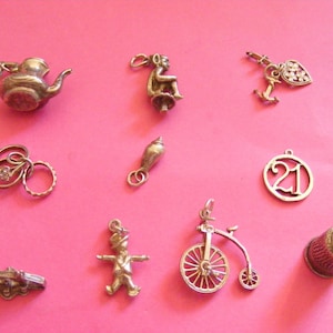 May include: A collection of ten silver-toned metal charms including a teapot, a seated figure, a heart with an anchor, a ring set, a seashell, a circular charm with the number 21, a horizontal bar, a standing figure, a bicycle, and a thimble.