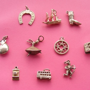 May include: A collection of silver charms on a pink background. Charms include a horseshoe, a sailboat, a cowboy boot, a teardrop, a ballerina, a decorative circle, a treasure chest, a camera, a double-decker bus, and a cupid.