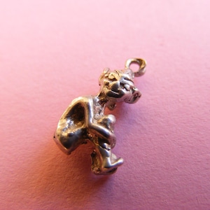 May include: A small, silver-toned charm depicting a figure holding another figure. The charm has a loop at the top for attachment. The figures have textured details, and the charm is set against a pink background.