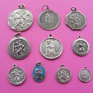 May include: A selection of silver Saint Christopher pendants in different shapes and sizes. The pendants feature Saint Christopher carrying the Christ child, with detailed relief work. One pendant has a blue enamel background. The pendants are arranged on a pink backdrop.