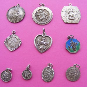 Könnte beinhalten: Eine Sammlung silberfarbener religiöser Anhänger und Charms, jeweils mit aufwendigen geprägten Designs. Die Anhänger variieren in der Form, darunter rund, herzförmig und achteckig, und zeigen religiöse Figuren und Symbole. Ein Anhänger hat blaue und grüne Emaille-Akzente.