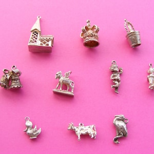 May include: A collection of silver charms, including a castle, crown, bucket, bells, donkey, two figures, a duck, a cow, and a lion. The charms are displayed against a bright pink background.