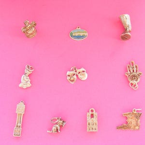 Könnte beinhalten: Eine Sammlung goldfarbener Charms auf pinkfarbenem Hintergrund. Die Charms zeigen eine Krone, ein ovales Landschaftsbild, einen Stiefel, eine Büste, Komödien- und Tragödienmasken, eine Hand mit einem Auge, eine Standuhr, einen Hund, ein Gebäude und eine Karte.