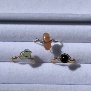 May include: Three gold wire wrapped rings with different stones. The first ring has a light pink oval stone, the second ring has a light green stone, and the third ring has a black round stone.