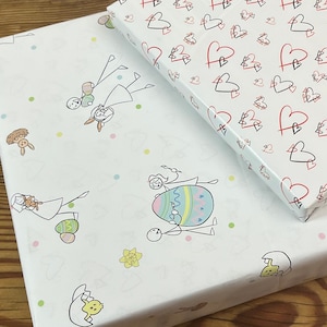 Könnte beinhalten: Zwei verpackte Geschenke auf einer Holzoberfläche. Eines ist mit weißem Papier mit roten und schwarzen Herzen bedeckt. Das andere ist in weißes Papier mit Osterillustrationen eingewickelt.